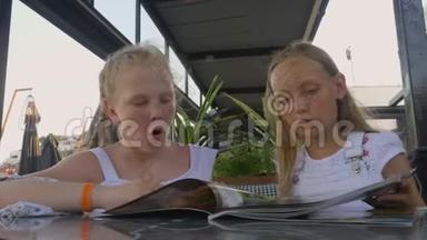 两个女孩在户外夏季咖啡馆寻找菜单。 年轻女孩青少年在餐厅菜单上交谈和选择食物。